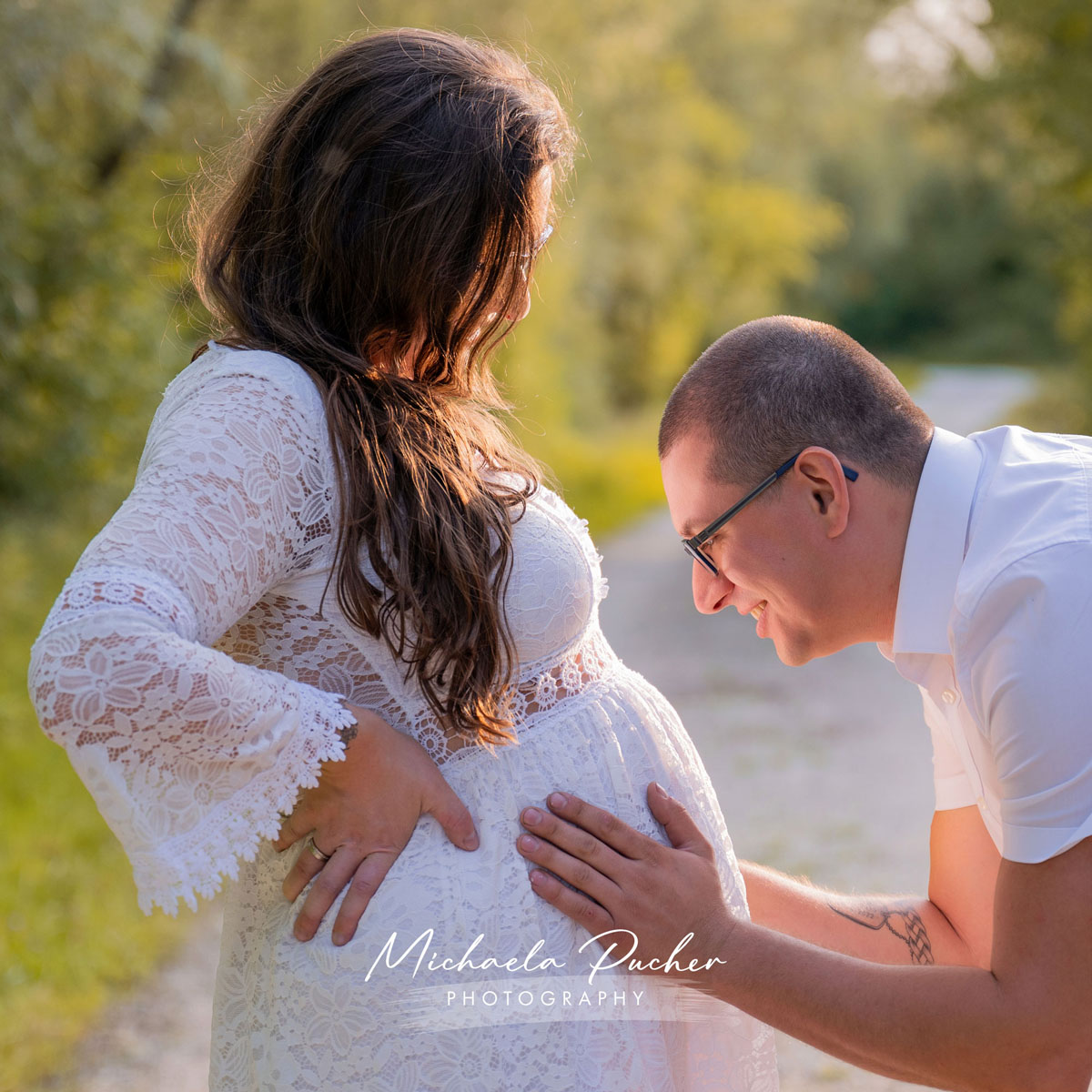 Partner kniet vor der schwangeren Frau, legt liebevoll die Hände auf ihren Babybauch und lächelt dabei voller Vorfreude; warmes Sonnenlicht umrahmt die Szene.